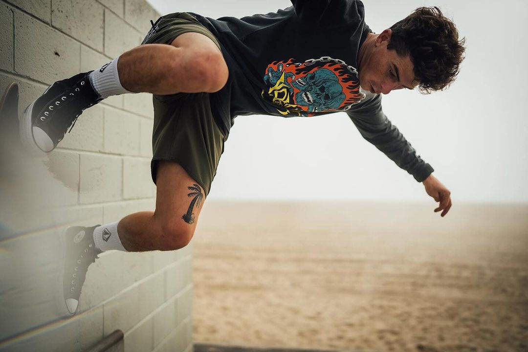 Athletic man in black long-sleeve shirt and green shorts doing a stunt on a beachside wall with sand and hazy sky in the background.