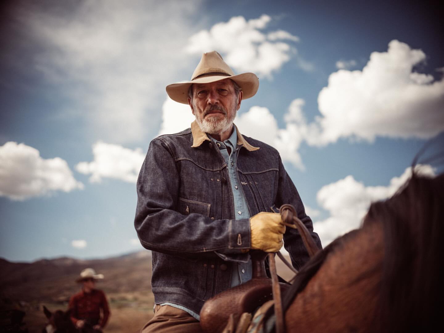 Portrait of a mature man in a denim jacket and cowboy hat on horseback against a cloudy sky, with another rider in the background.