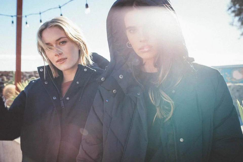 Two women wearing black puffer jackets stand outside on a sunny day. The blonde woman is to the left, the brunette woman is to the right.