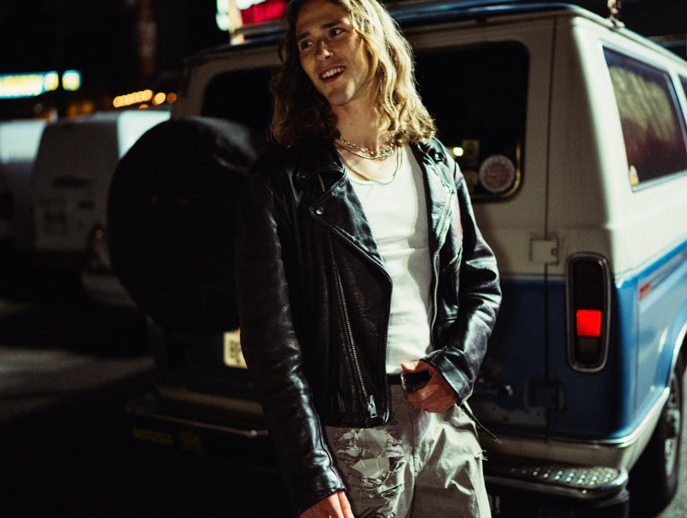 Man with long hair wearing a leather jacket poses near a vintage blue and white van at night with city lights in the background.