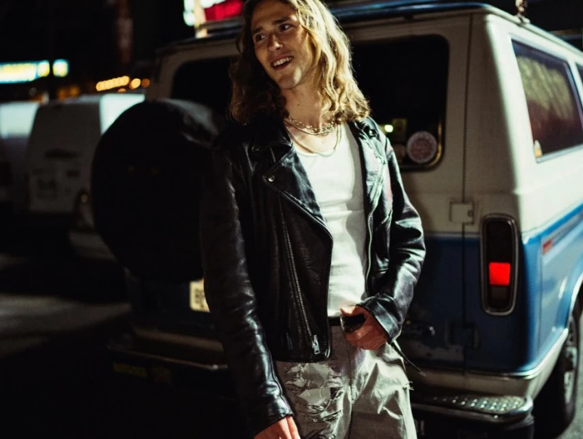 Man with long hair wearing a leather jacket poses near a vintage blue and white van at night with city lights in the background.