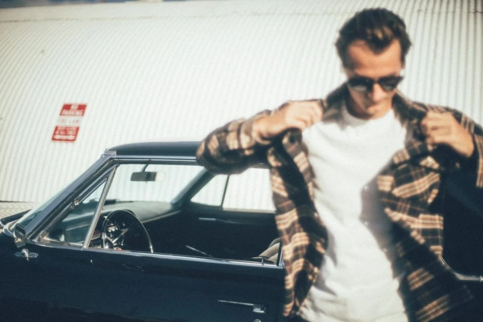 Man wearing sunglasses and a plaid shirt stands near a classic black car in front of a corrugated metal building, creating a stylish, vintage scene.