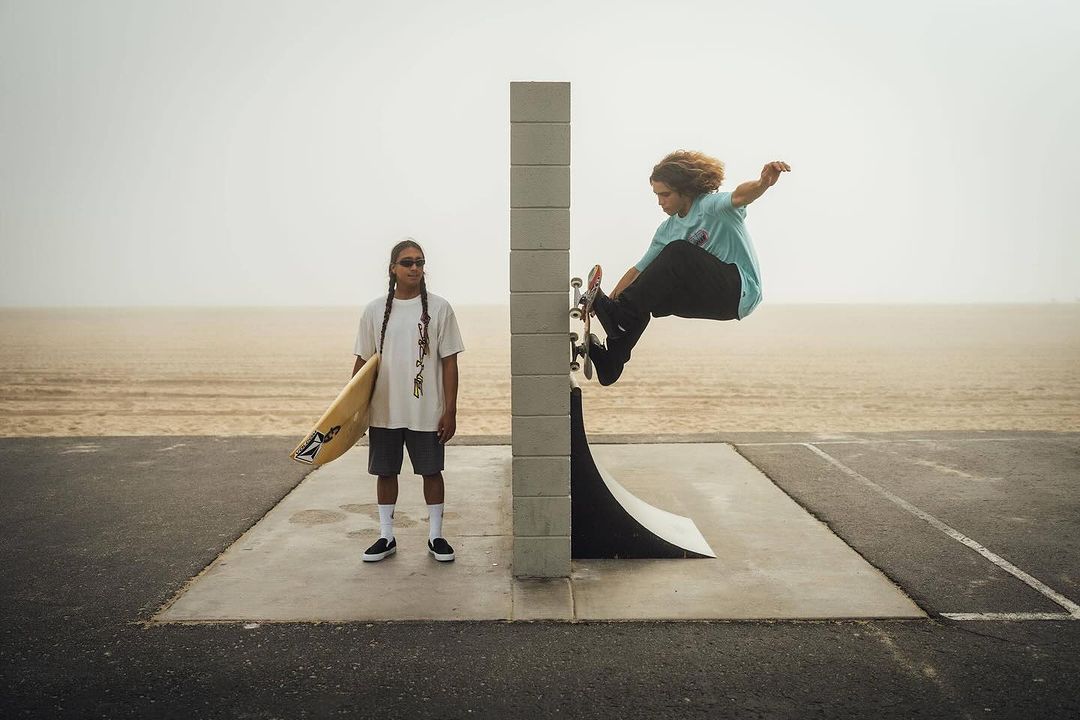 Two skateboarders at a beach skate park. One is in mid-air performing a trick, while the other watches, holding a surfboard.