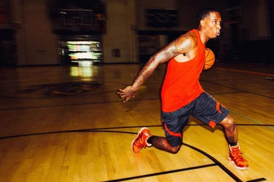 Black basketball player in red tank top and dark shorts dribbling a basketball on an indoor court.