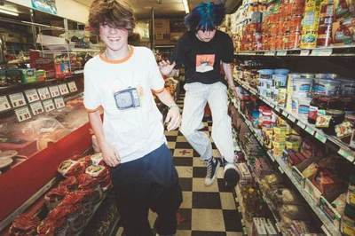 Two youths in a grocery store aisle with packed shelves, one smiling and one jumping mid-air.