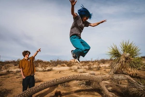 A person with blue hair leaps mid-air over a fallen Joshua tree branch in a desert landscape while another person points.