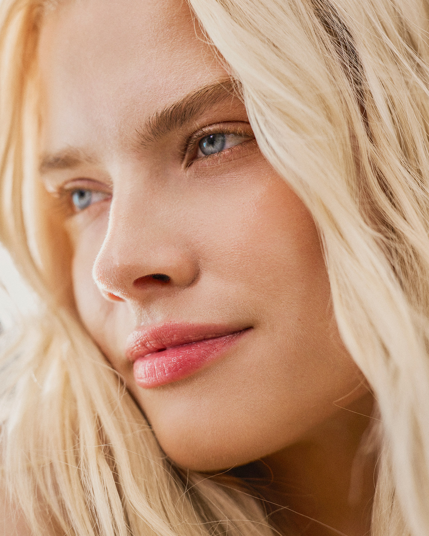 Close-up portrait of a fair-skinned adult female with blonde wavy hair and blue eyes, looking off-camera in a beauty-style shot.
