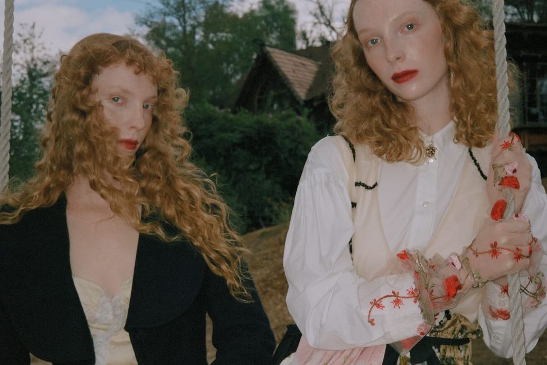 Two red-haired women pose outdoors wearing vintage-style clothing. One sits on a swing; a house and trees form the background.