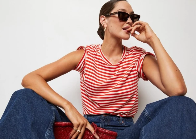 Model wearing dark sunglasses, red and white striped t-shirt, and wide-leg blue jeans holding a red woven bag against a white background.