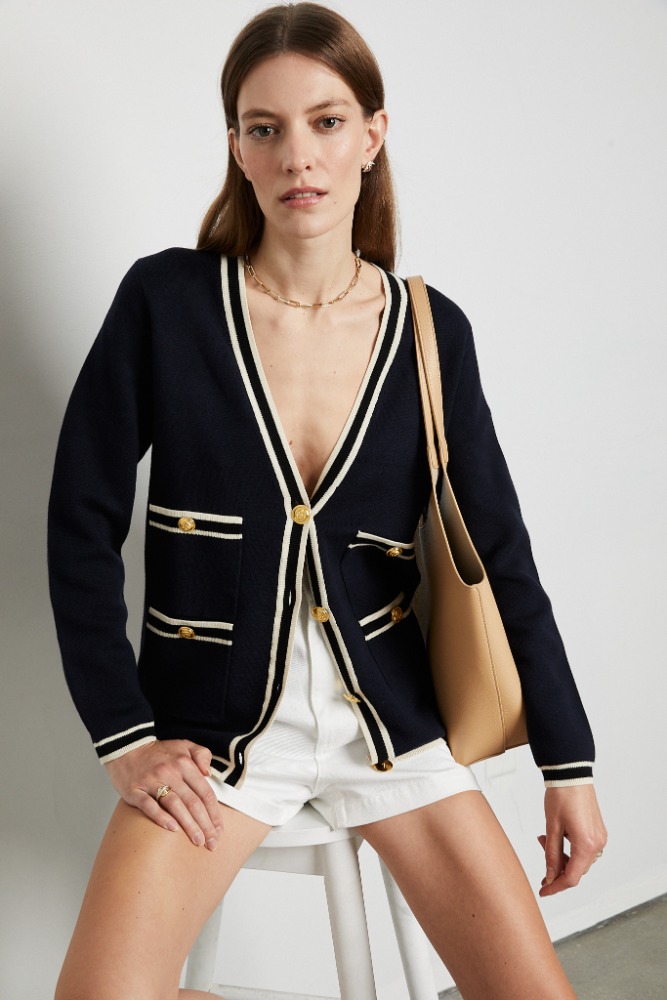 Fashionable woman poses in navy cardigan with white trim, gold buttons, white shorts, and a tan tote bag, seated indoors.