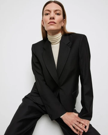 Woman wearing a black suit jacket and cream turtleneck sweater against a clean white background