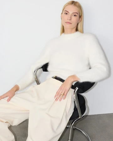 Blonde woman wearing a fluffy white sweater and cream trousers seated on a chrome chair against a light background.