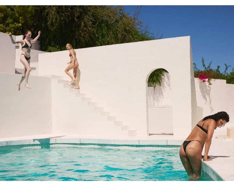 Three women in bikinis near a bright blue pool with white walls, lush trees, and clear sky creating a vibrant, summery scene.