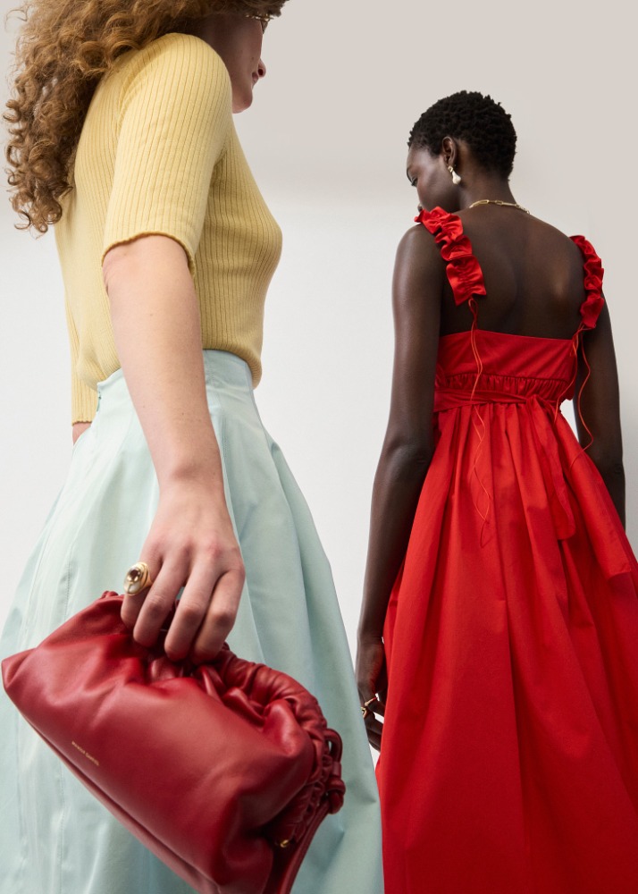Two women in dresses posing indoors. One woman is holding a red clutch, while the other wears a red dress. Fashion shot.