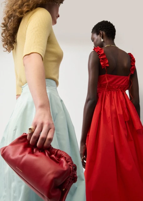 Two women in dresses posing indoors. One woman is holding a red clutch, while the other wears a red dress. Fashion shot.