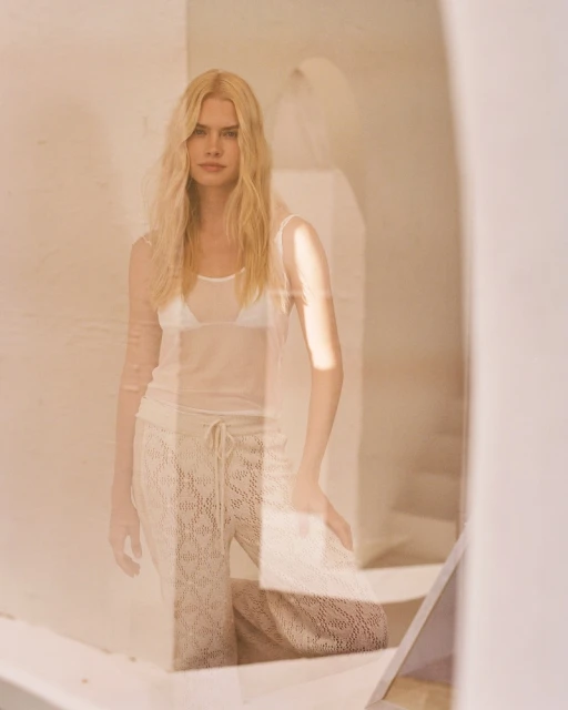 Woman with long blonde hair poses in sheer top and open-weave pants in an indoor setting with natural light and architectural details.
