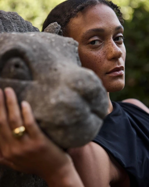 Close-up of an adult woman with dark hair and freckles posing next to a stone dragon head, wearing black clothing, outdoors, soft lighting.