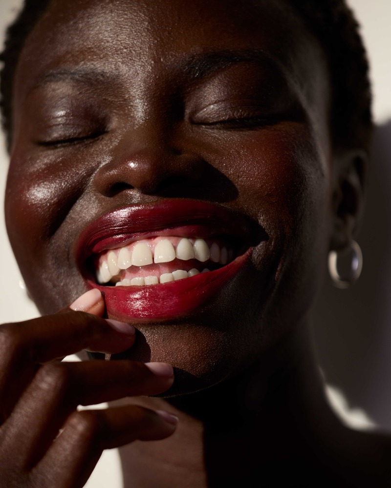 Close-up portrait of a smiling Black woman with dark glossy lipstick touching her lower lip by Ari Abramczyk.