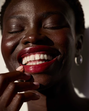 Close-up portrait of a smiling Black woman with dark glossy lipstick touching her lower lip by Ari Abramczyk.