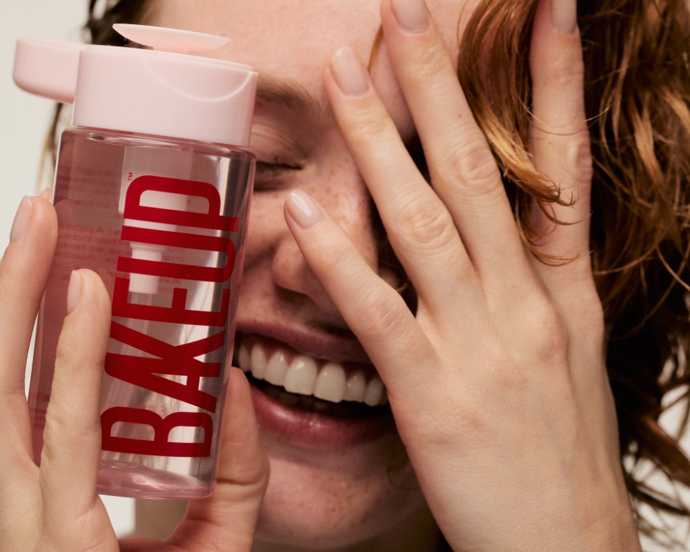 Woman smiling, holding makeup remover bottle with pink cap and red text. Close-up shot in beauty context.