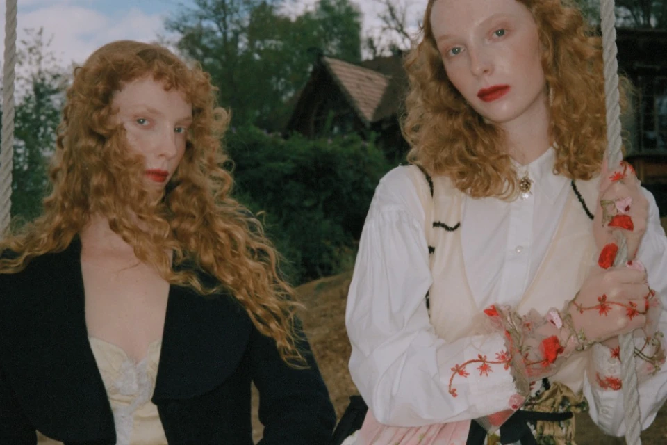 Two red-haired adult females pose outdoors in vintage fashion near a wooden house, with one on a flower-decorated swing.