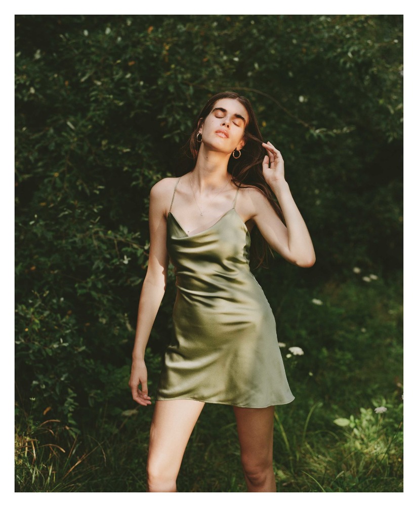 Woman with long brown hair poses in an olive green satin dress outdoors in a garden with blurred green foliage background, eye-level angle.