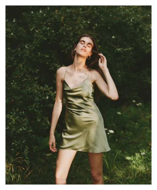 Woman with long brown hair poses in an olive green satin dress outdoors in a garden with blurred green foliage background, eye-level angle.