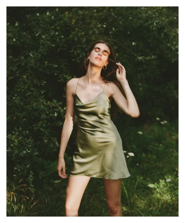 Woman with long brown hair poses in an olive green satin dress outdoors in a garden with blurred green foliage background, eye-level angle.