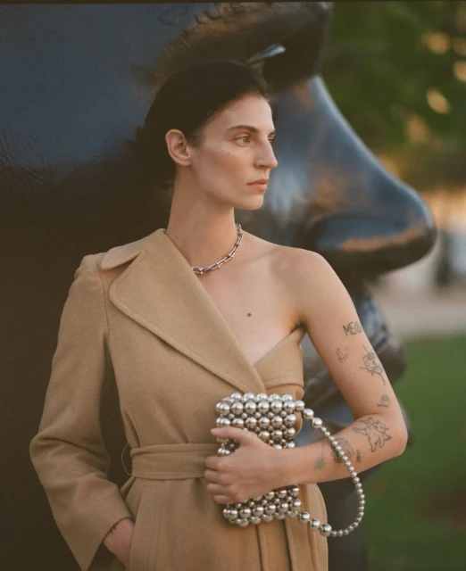A woman in a tan coat holds a silver beaded purse next to a large sculpture, blending fashion with art in an outdoor setting.