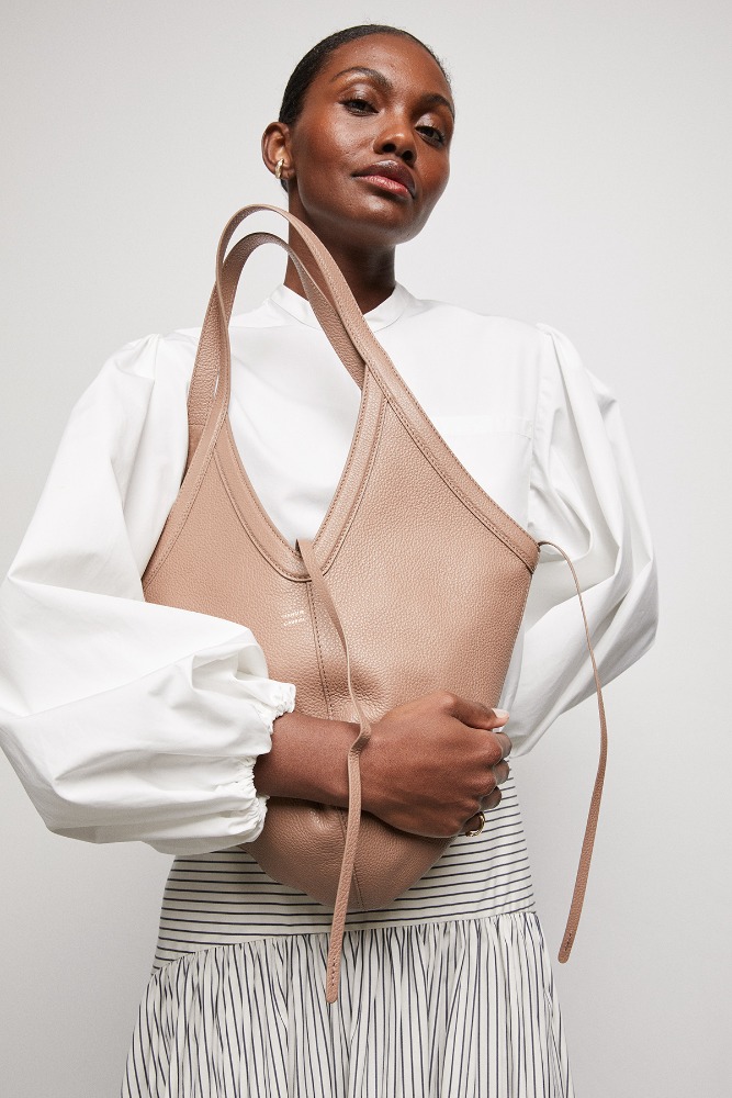 Studio shot of a woman posing with a light brown leather hobo bag, styled with a white blouse and striped skirt against a neutral backdrop.