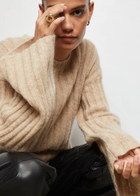 Fashion shot of an adult female wearing a beige ribbed sweater with flared sleeves and black pants, accessorized with a golden ring.