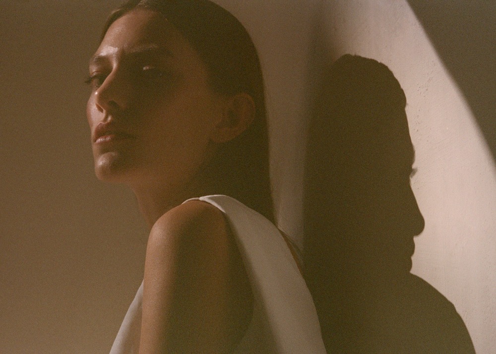 Close-up studio portrait of an adult woman with tan skin in white top, facing away from the camera, with dramatic lighting and shadow.