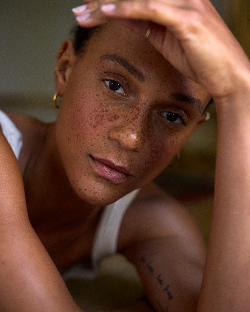 Close-up portrait of a woman with prominent freckles and natural makeup, raising her hand to her forehead.