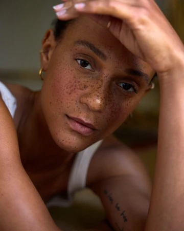 Close-up portrait of a woman with prominent freckles and natural makeup, raising her hand to her forehead.