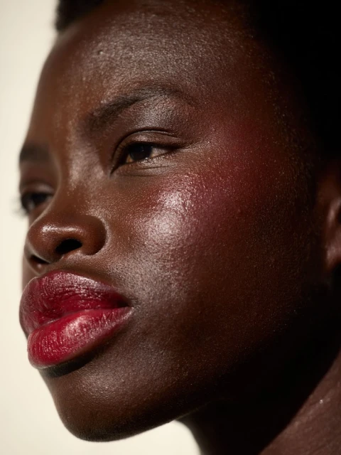 Close-up portrait of a Black woman with dark brown skin, red lipstick, and a glossy complexion against a neutral-toned background.
