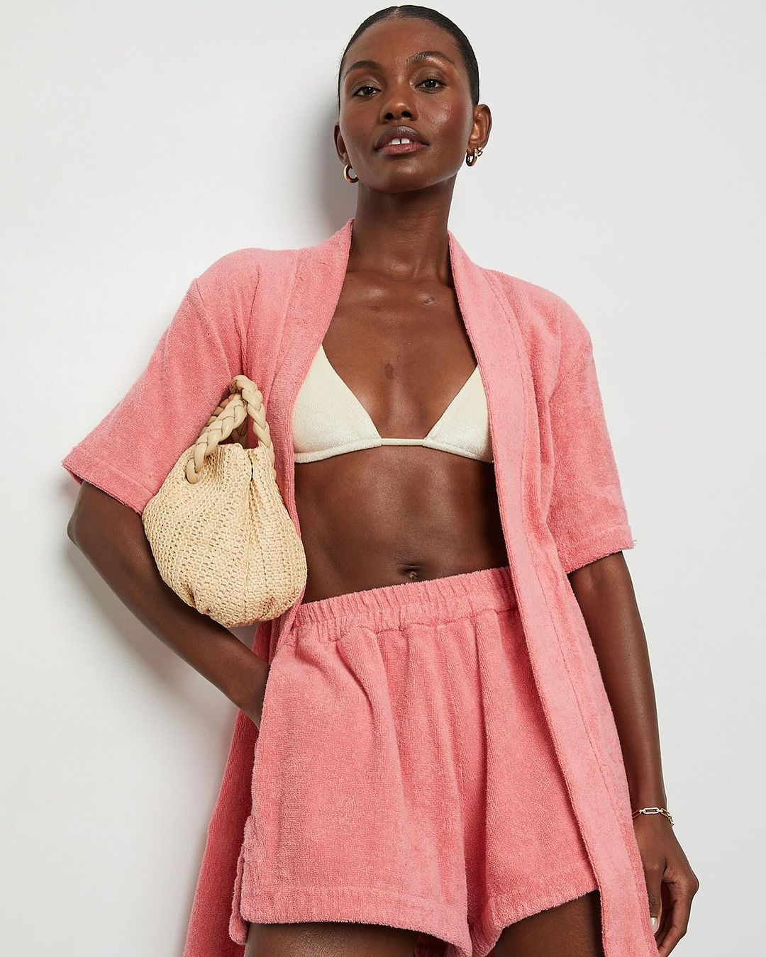 Woman in pink terry cloth shorts and robe, holding a woven handbag, posing against a white background in a studio shot.