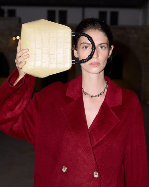 Adult female in a red coat holding a yellow geometric purse with a black handle and wearing a silver necklace outdoors.