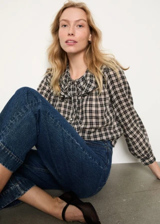 A seated woman wearing a plaid ruffle collar blouse and blue denim jeans against a neutral grey studio background.