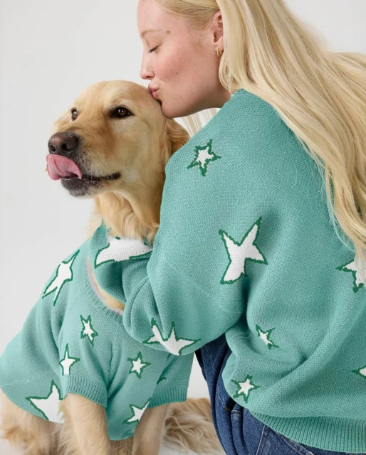 Adult female kisses Golden Retriever dog wearing matching star-patterned sweater against white background in an indoor setting.