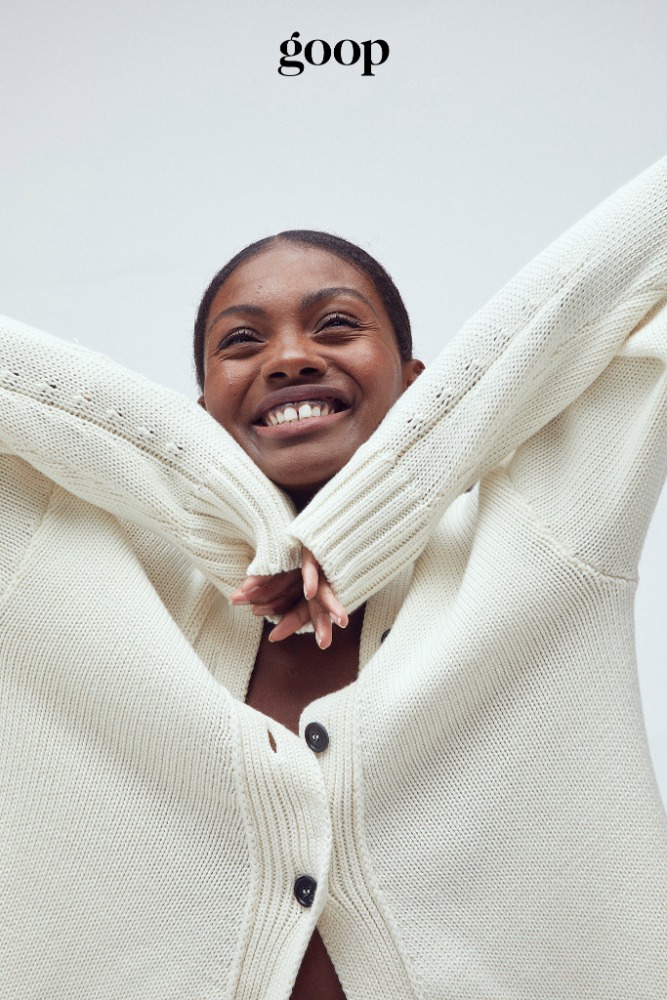 Smiling dark-skinned woman in a cream-colored button-down sweater with her arms up, in a close-up studio shot against a neutral backdrop.