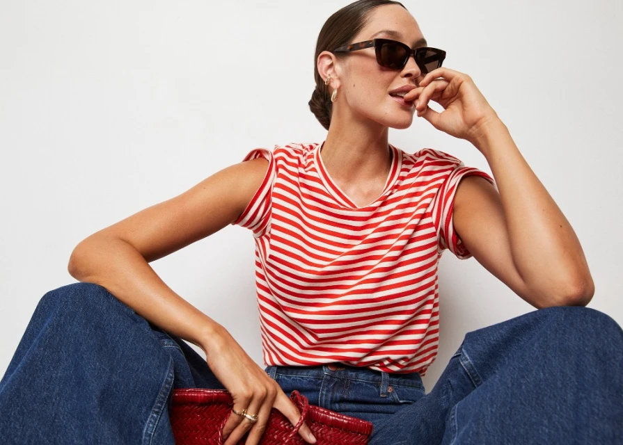 Adult female poses seated in blue denim with a red and white striped top, sunglasses, a red woven bag, and jewelry indoors.