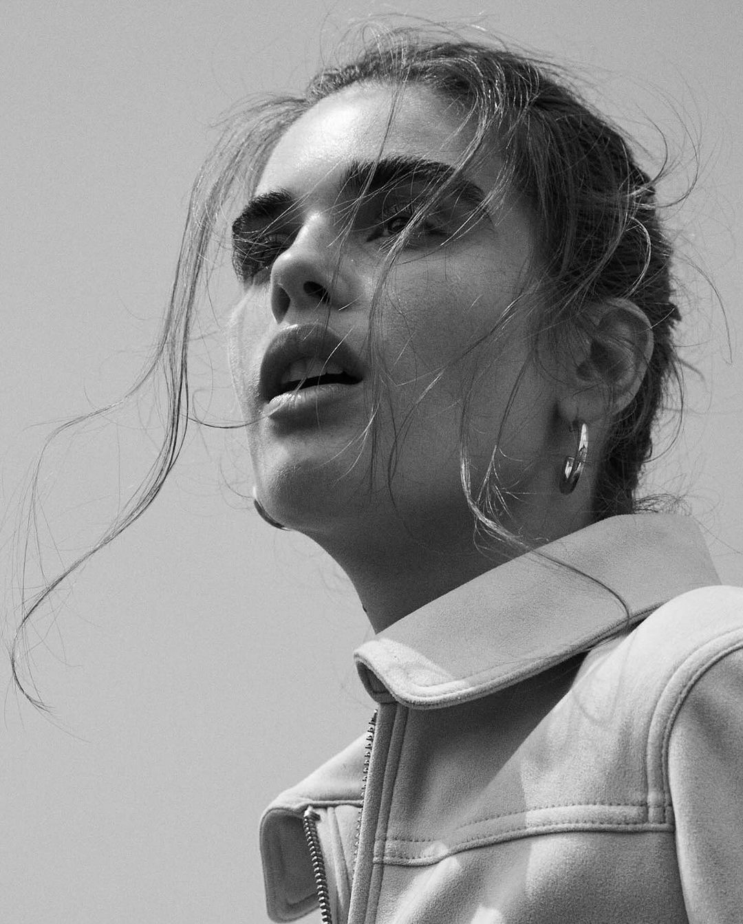 Monochromatic portrait of a woman looking upwards, wearing a jacket and hoop earrings against a bright sky, with windblown hair.