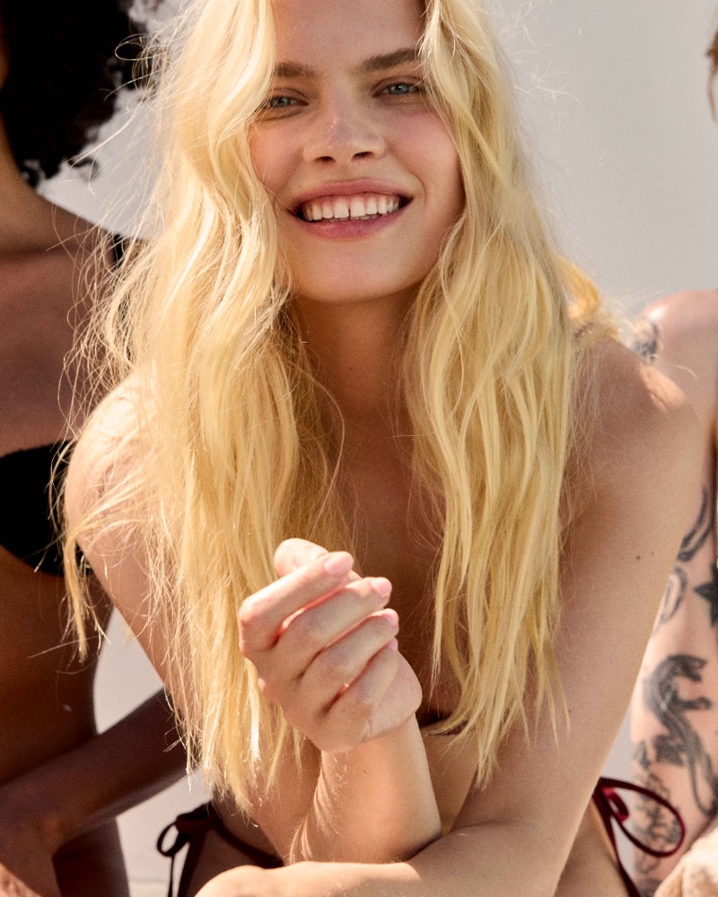 Close-up of a smiling blonde woman, outdoors, wearing a bikini top, with other women partially visible in the background.