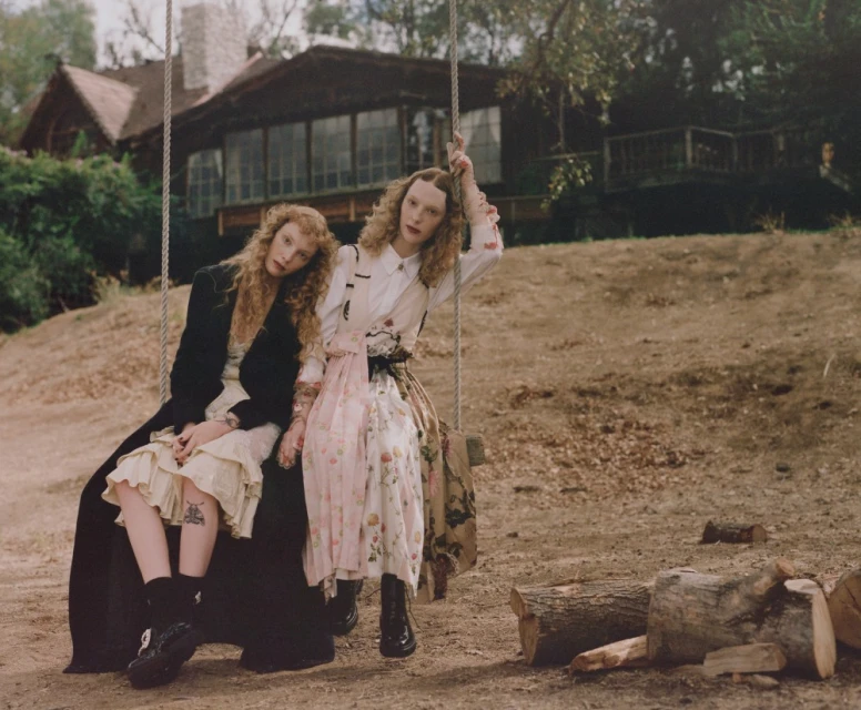 Two women sit on a rope swing in dresses and dark jackets with a rural home visible in the background during daytime.