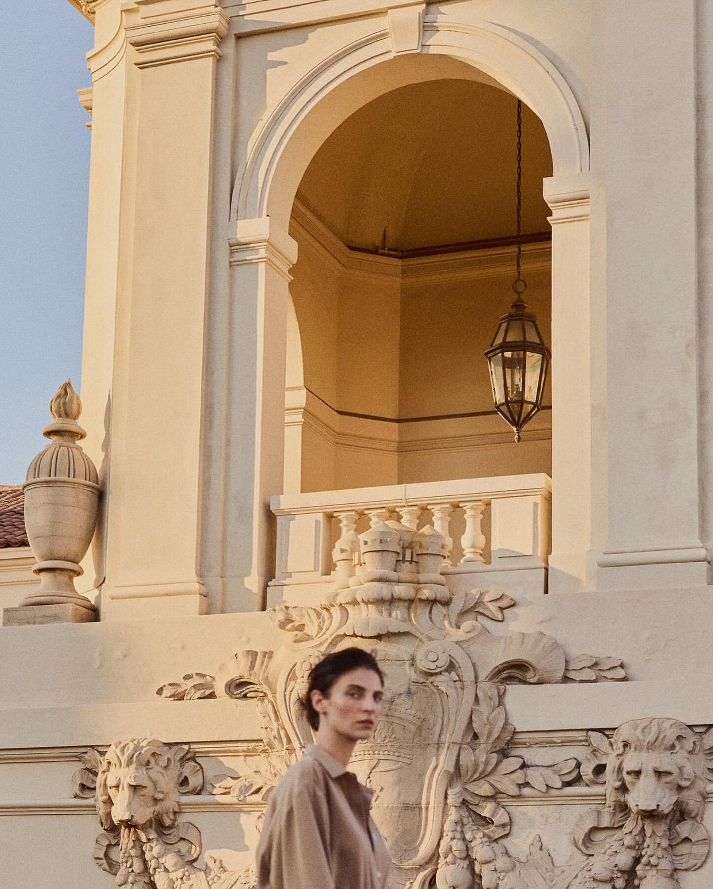 Woman in casual dress stands against decorative architecture with arches, an ornate light fixture, and stone lions in the background.