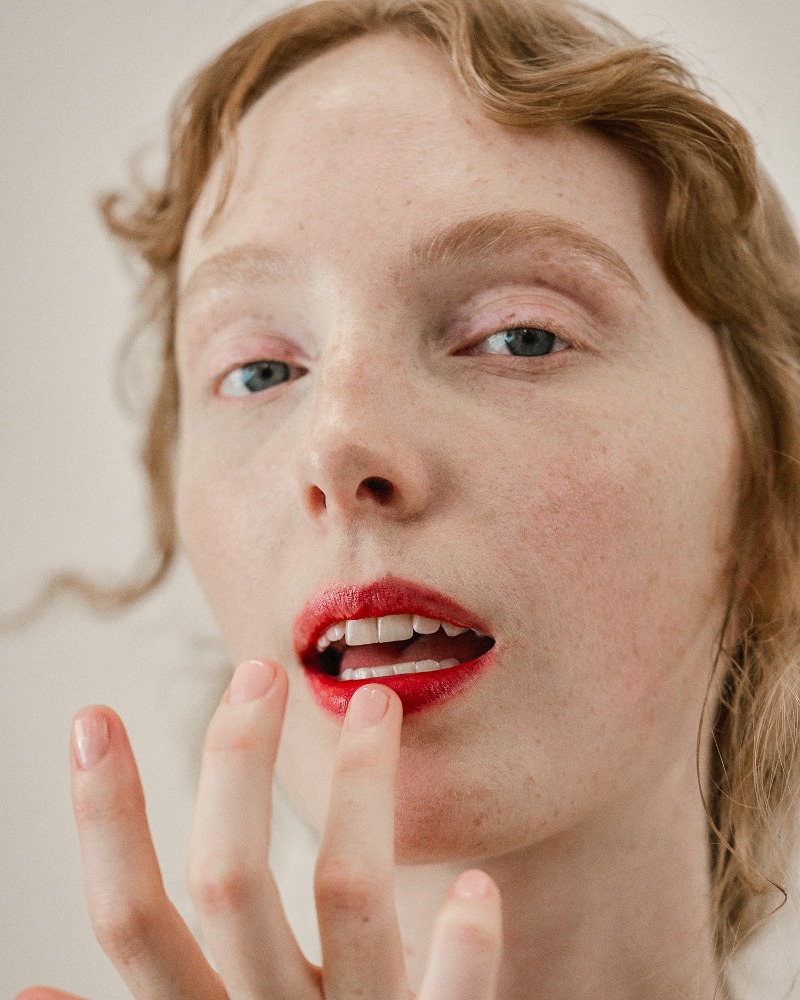 Close-up portrait of an adult female with red lipstick touching her lip with her fingers, soft studio lighting, and neutral backdrop.
