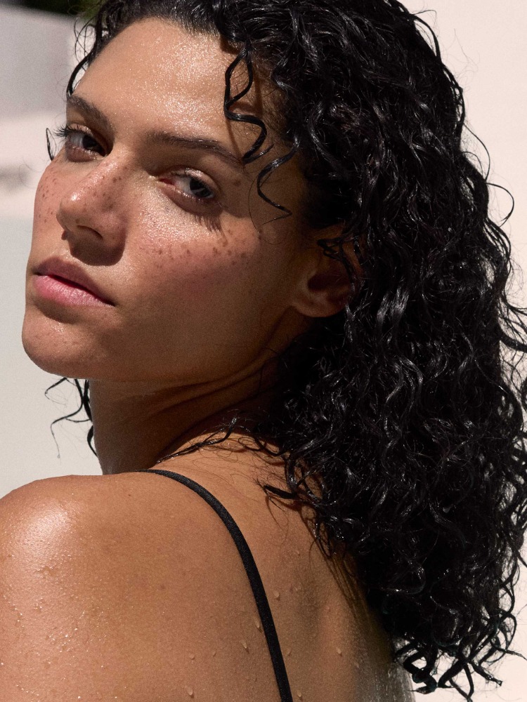 Close-up portrait of a woman with wet dark curly hair, freckles, and a black strap top against a bright background.