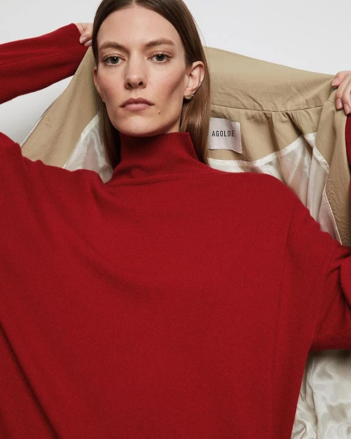 A woman with fair skin and medium-length brown hair wears a red turtleneck sweater while holding a beige coat, posing indoors.