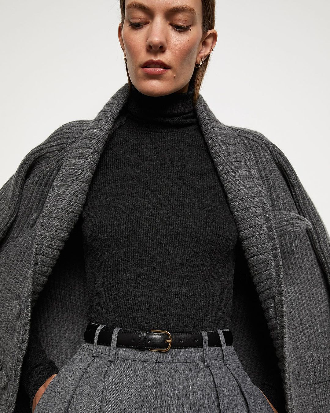 A sophisticated fashion image featuring an adult woman in a dark turtleneck, grey trousers, and a stylish grey cardigan against a plain background.