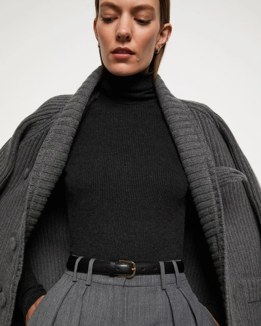 A sophisticated fashion image featuring an adult woman in a dark turtleneck, grey trousers, and a stylish grey cardigan against a plain background.
